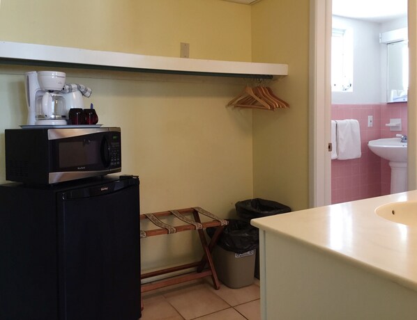 Una cocina compacta con microondas, cafetera y un refrigerador pequeño. Se observa un perchero de madera con perchas y un cesto de basura.