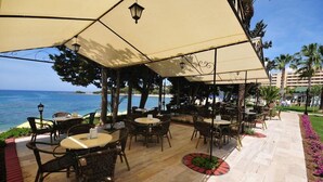 An outdoor seating area with tables and chairs under large umbrellas, overlooking a beach and ocean. - Incekum Su Hotel (Alanya)
