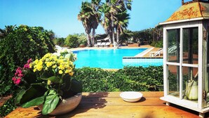 Una terraza de madera con una maceta, un farol y vistas a una piscina con palmeras al fondo.