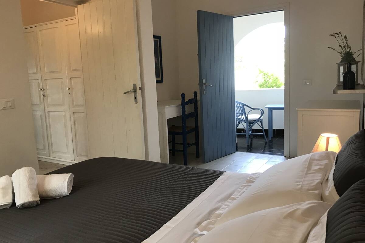 A hotel room with a bed, a desk, and a chair. There is a door leading to another room and a window with a view of greenery outside.