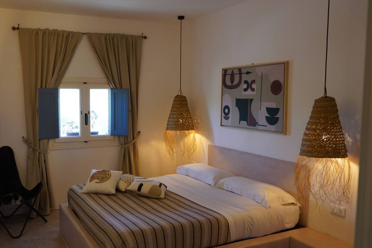 A bedroom with a bed, two hanging lamps, a desk with a chair, a window with curtains, and a wall with a piece of abstract art.