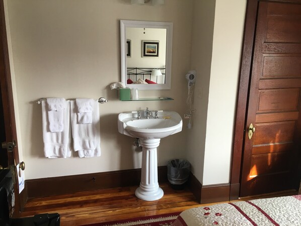 A bathroom with a pedestal sink, a mirror, a towel rack with white towels, a hairdryer, and a wooden door. - The Rebekah Inn (Old Orchard Beach)