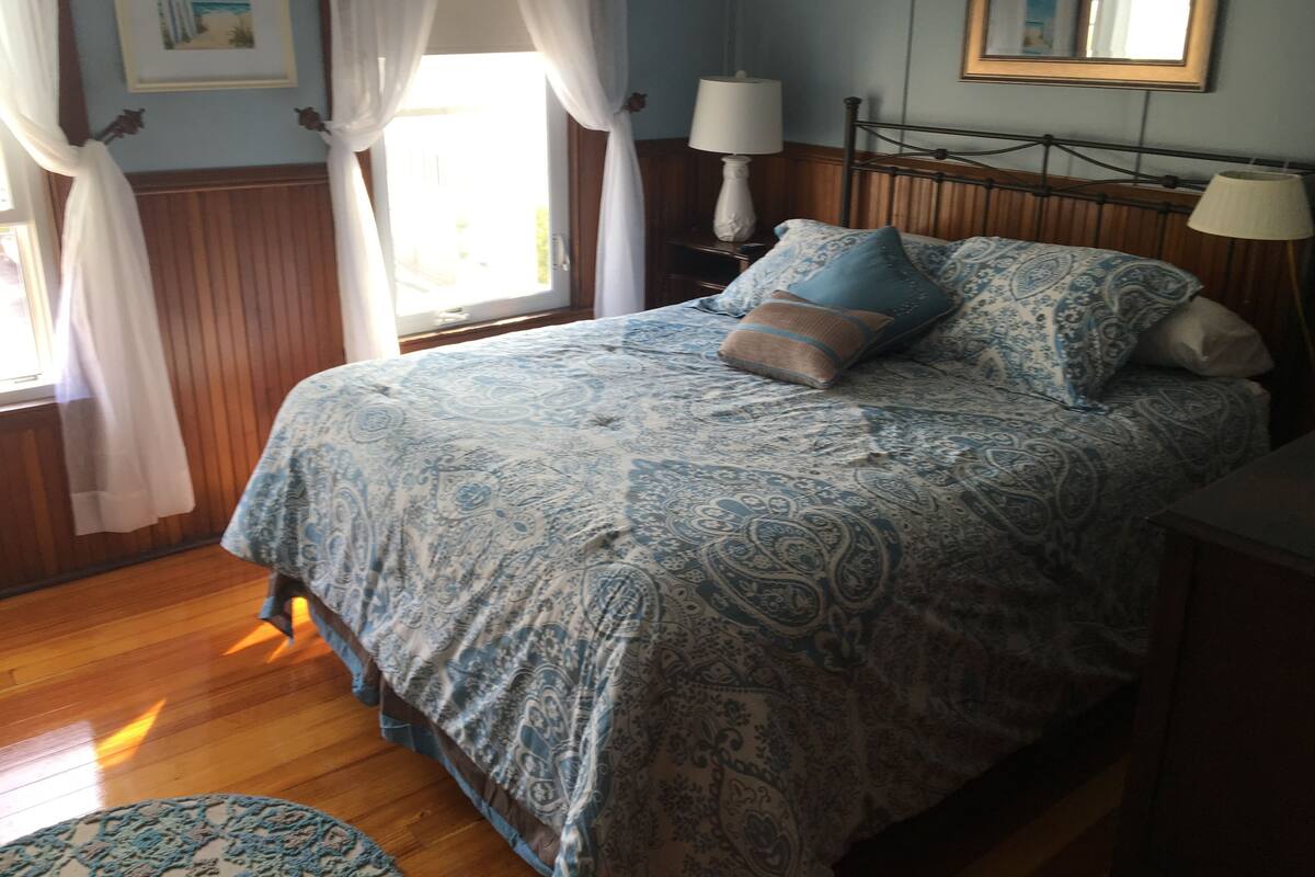 Un dormitorio con cama, dos lámparas de noche, una mesita de noche, una ventana con cortinas y un cuadro enmarcado en la pared.