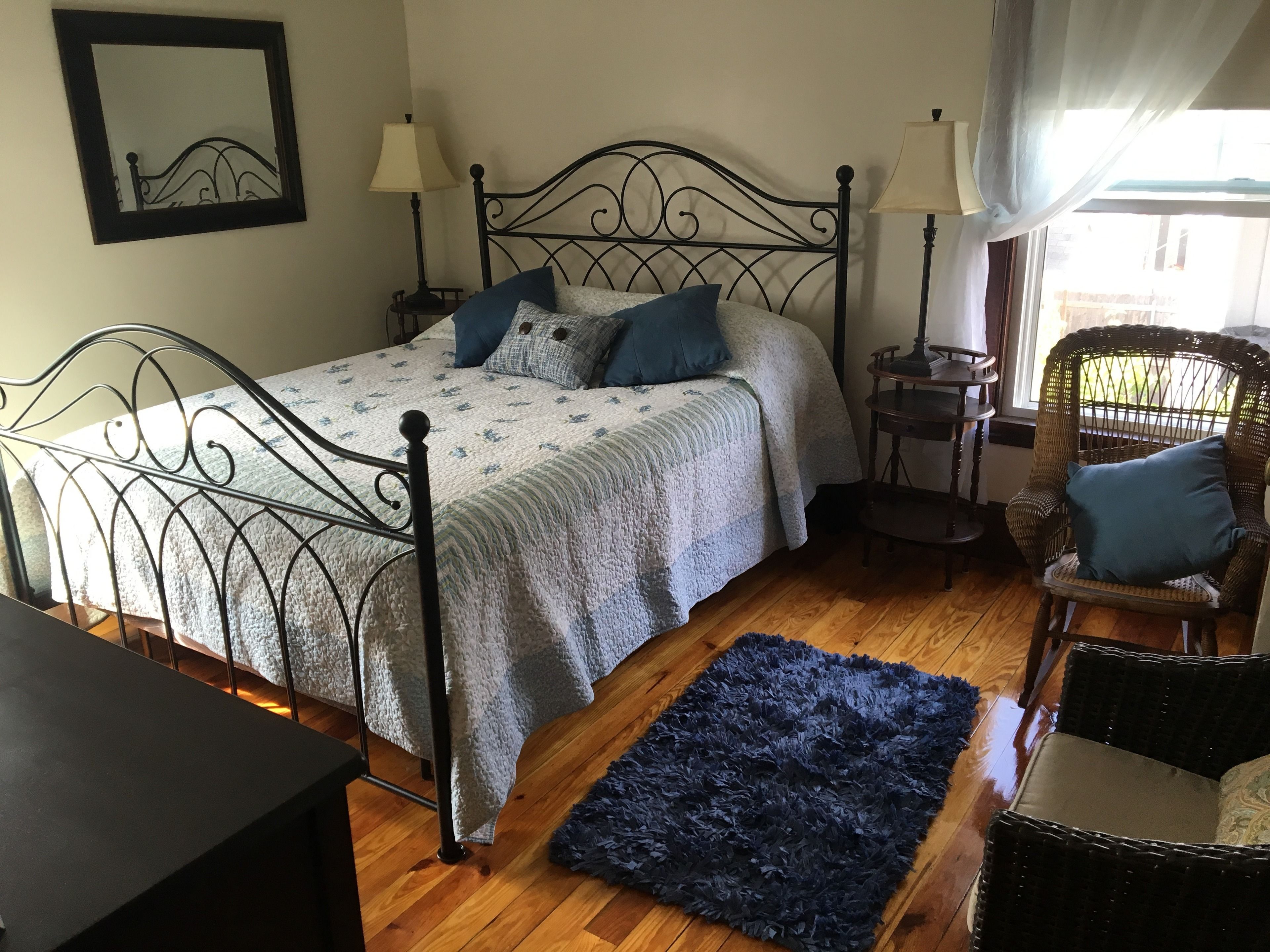 A bedroom with a metal bed frame, a large bed with multiple pillows, a dark blue rug, a wicker chair, and a small table with a lamp.