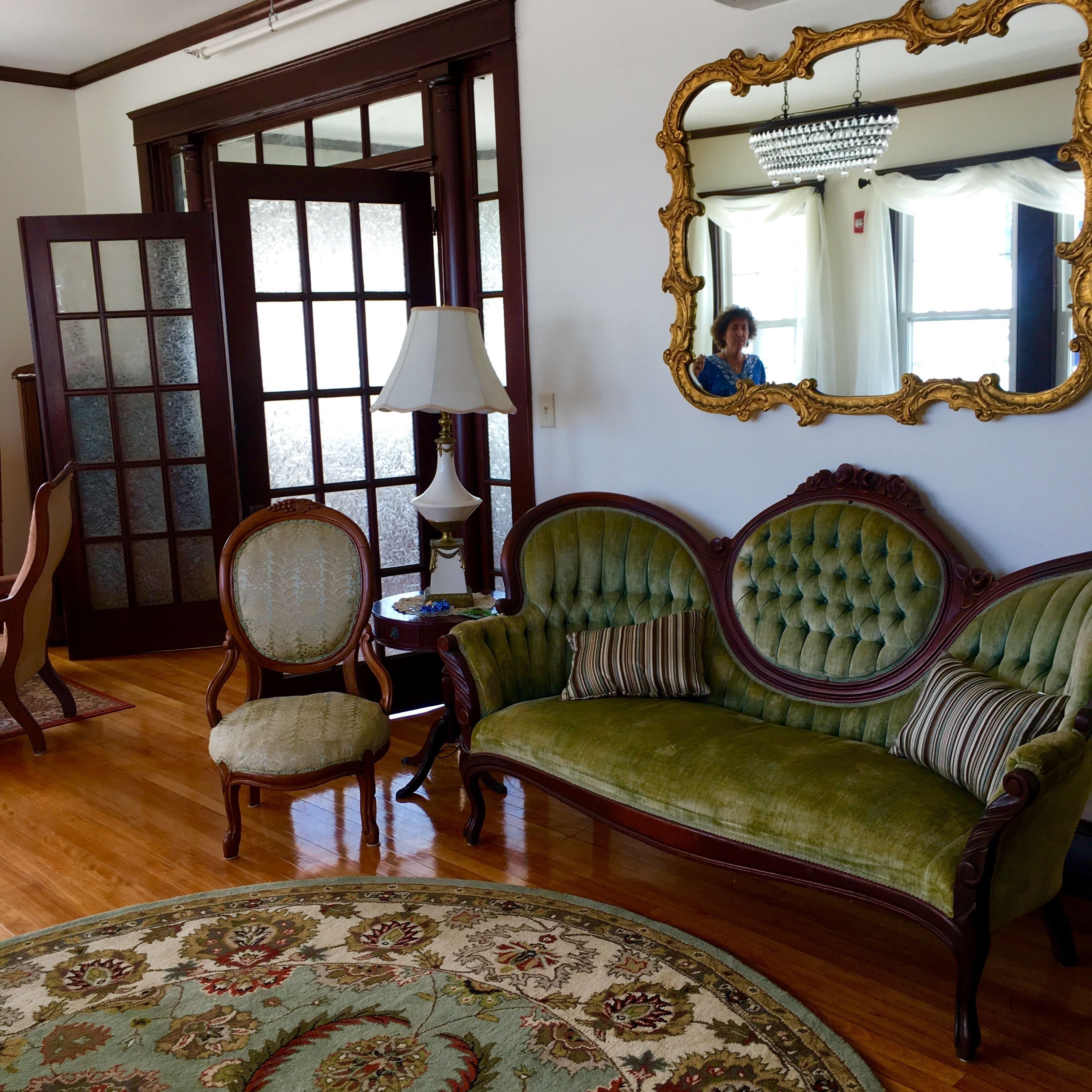 A room with a green sofa, a wooden chair, a round table, and a large mirror with an ornate frame.