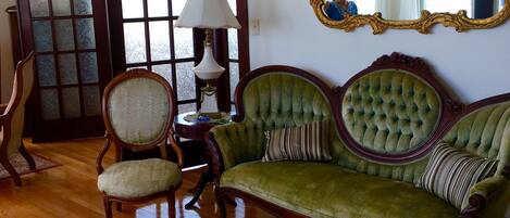 Una habitación con un sofá verde, una silla de madera, una mesa redonda y un espejo grande con un marco ornamentado.