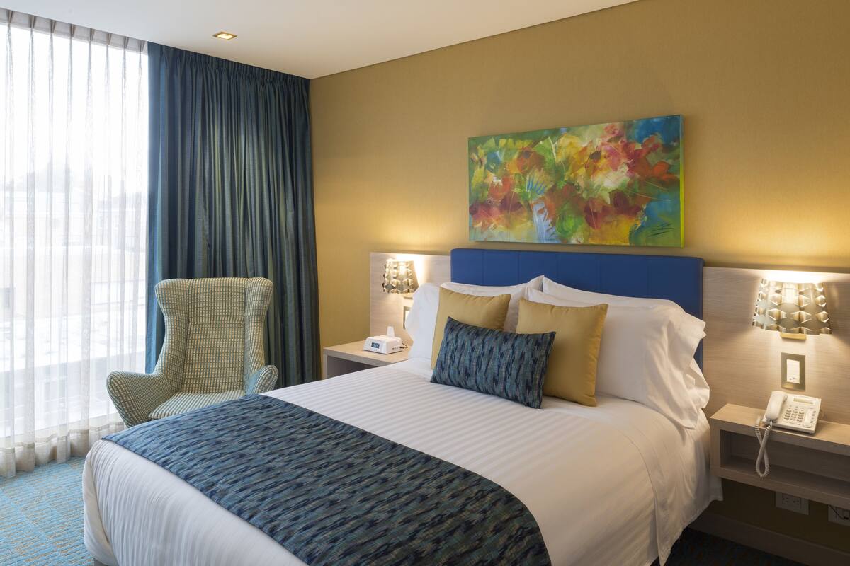 A hotel room with a large bed, a chair, a bedside table with a lamp, and a painting on the wall.
