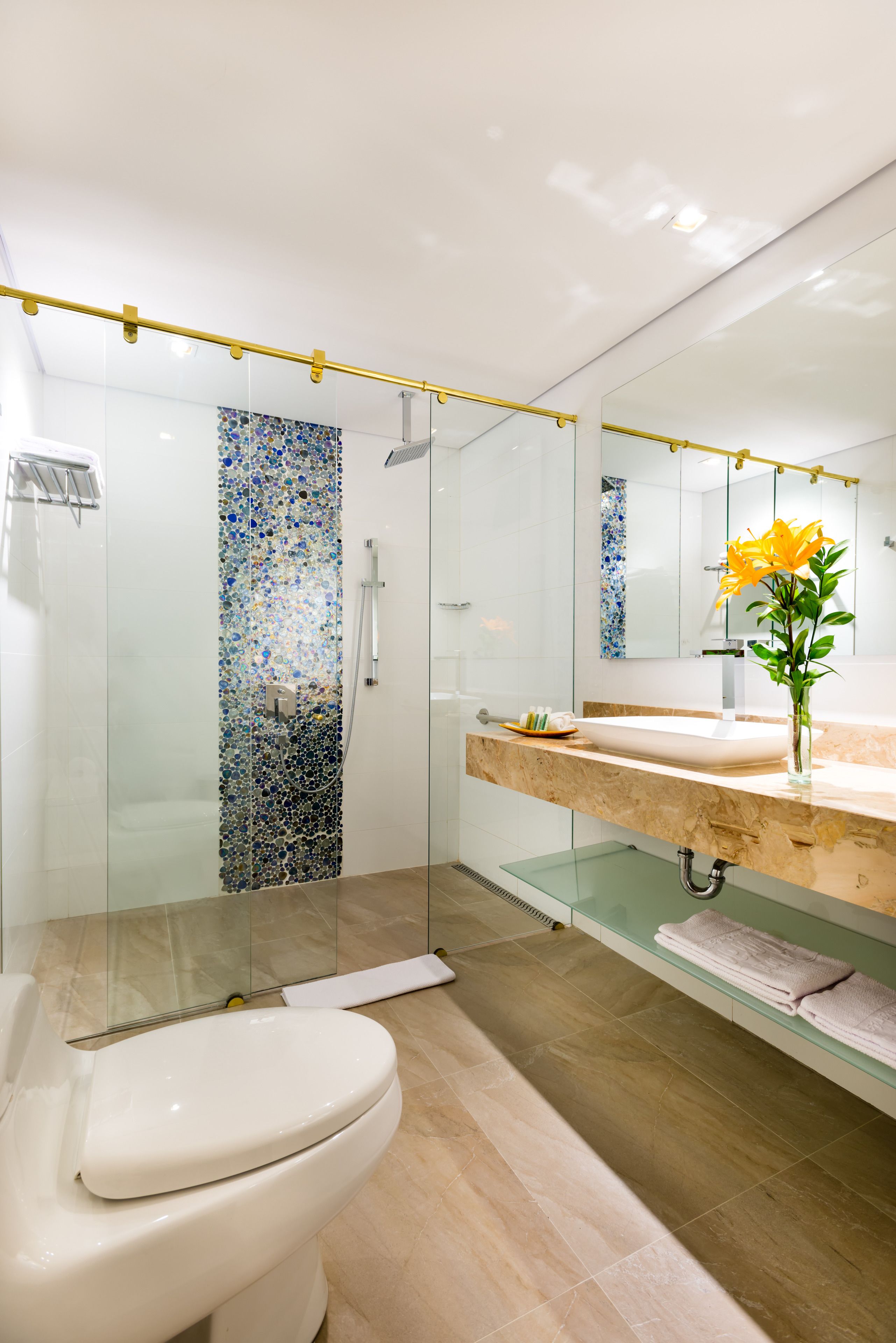 Une salle de bain moderne avec une douche en verre, des toilettes blanches, un lavabo en marbre et un miroir agrémenté d’un vase de fleurs.