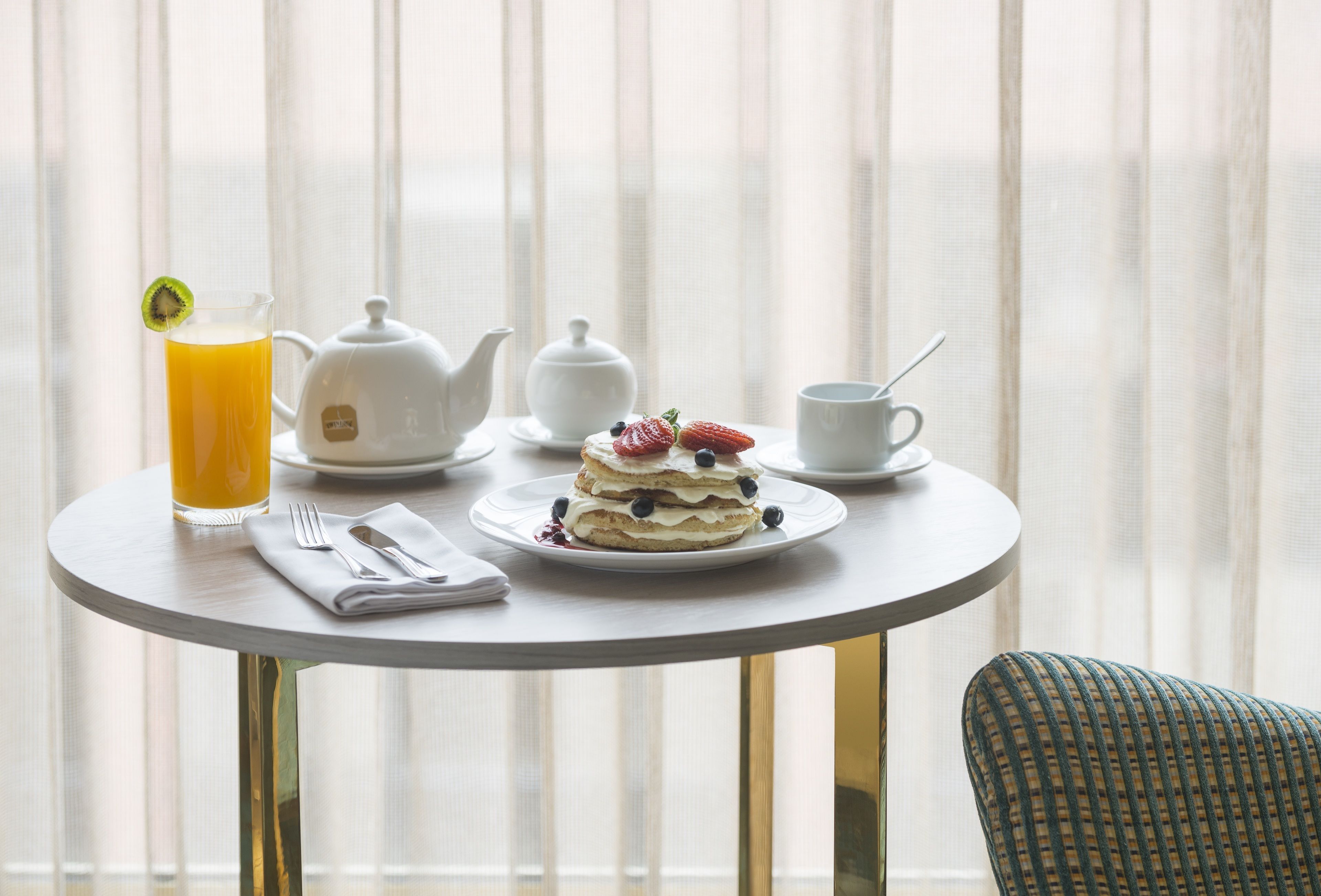 A breakfast table with a glass of orange juice, a teapot, a cup, a plate with a dessert, and a chair.