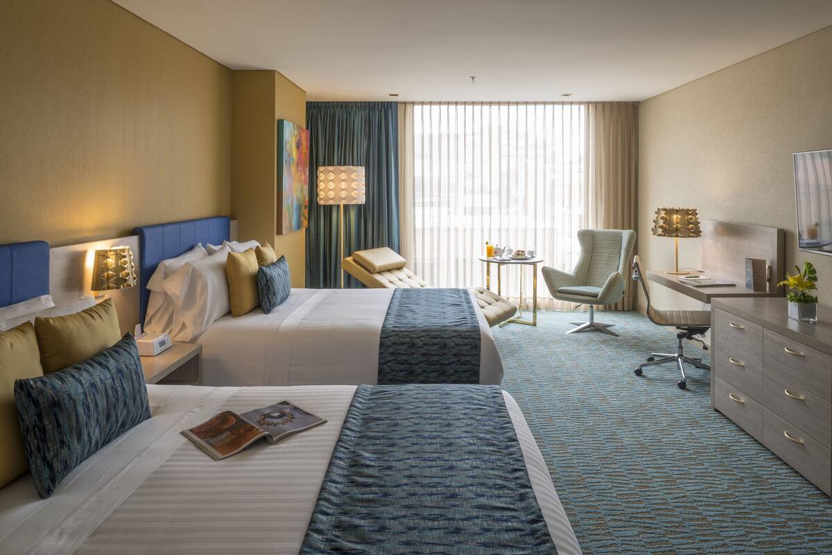A hotel room with two beds, a desk, a chair, and a large window with curtains.