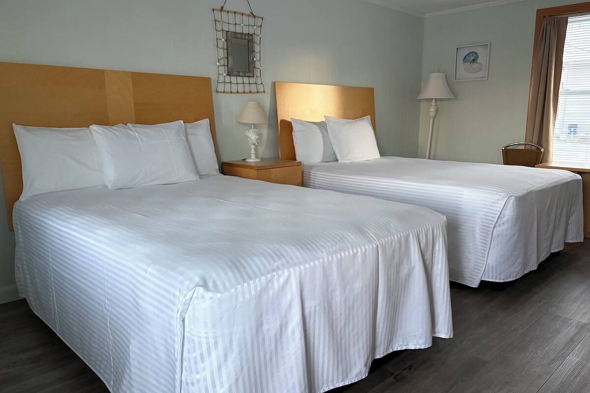 A hotel room with two beds, wooden headboards, white bedding, a bedside table with a lamp, a wall-mounted mirror, and a window with curtains.