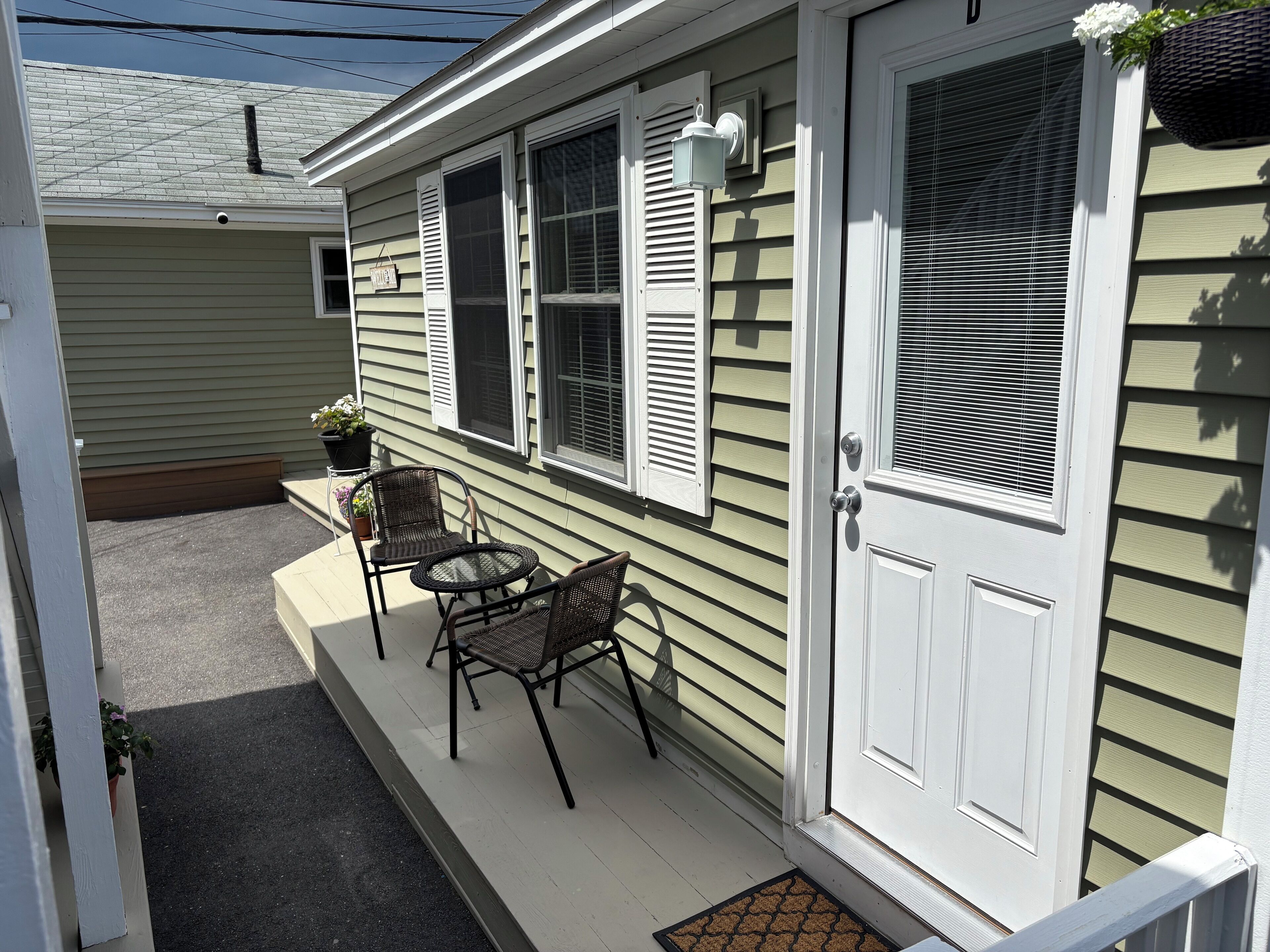 A small porch with two wicker chairs and a table, a white door, and a window with shutters.
