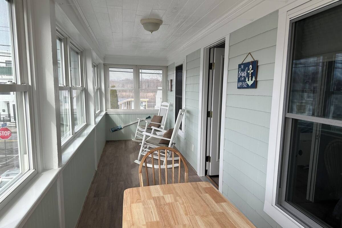 A narrow corridor with white walls and wooden flooring, leading to a door. There are several rocking chairs and a wooden table along the corridor.
