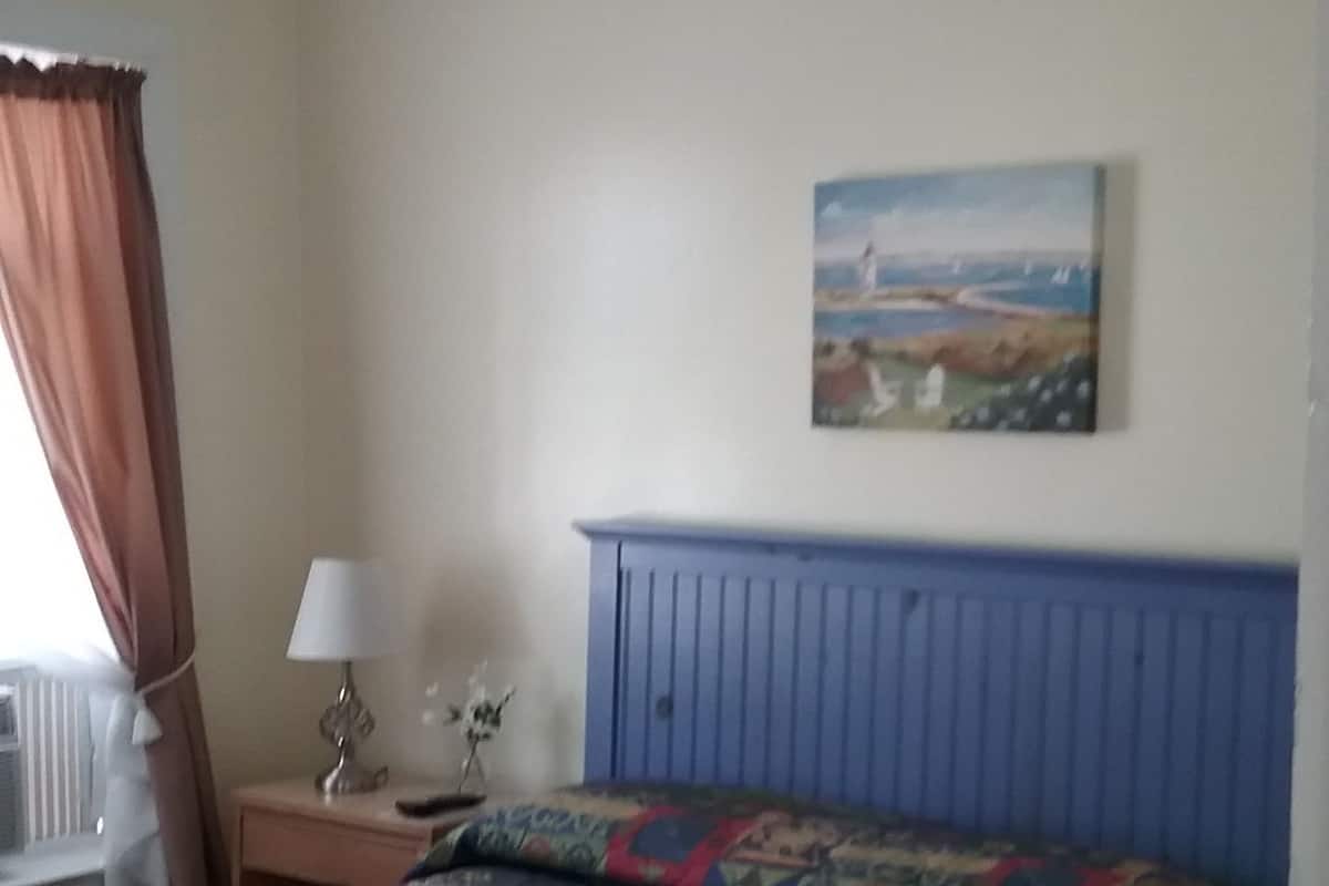 A bedroom with a bed, a nightstand, a lamp, a painting, and a window with curtains.