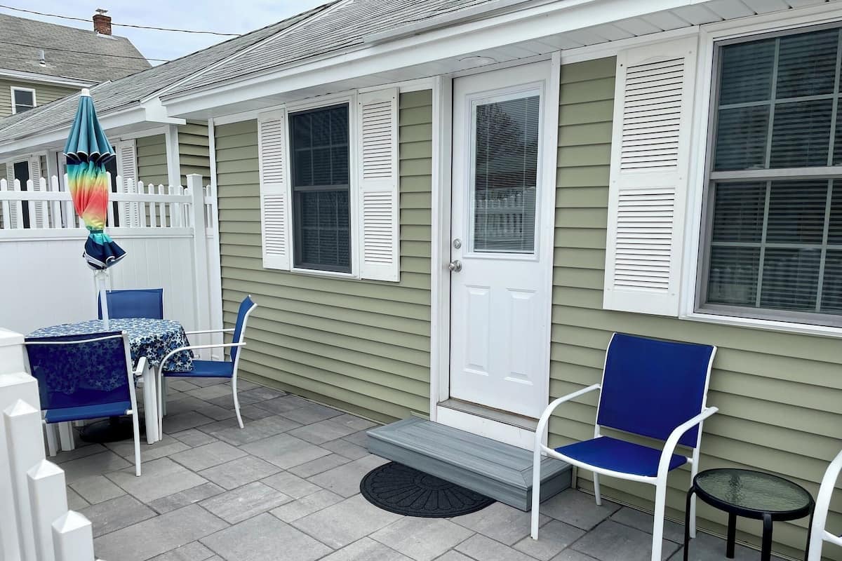A covered patio with a white door, two blue chairs, a small table, and a colorful umbrella.