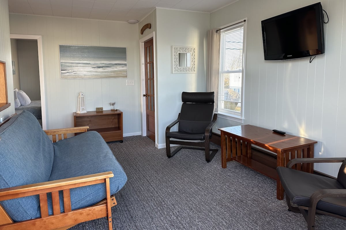 A cozy room with a wooden sofa, a black chair, a small table, a TV mounted on the wall, and a painting of a seascape.