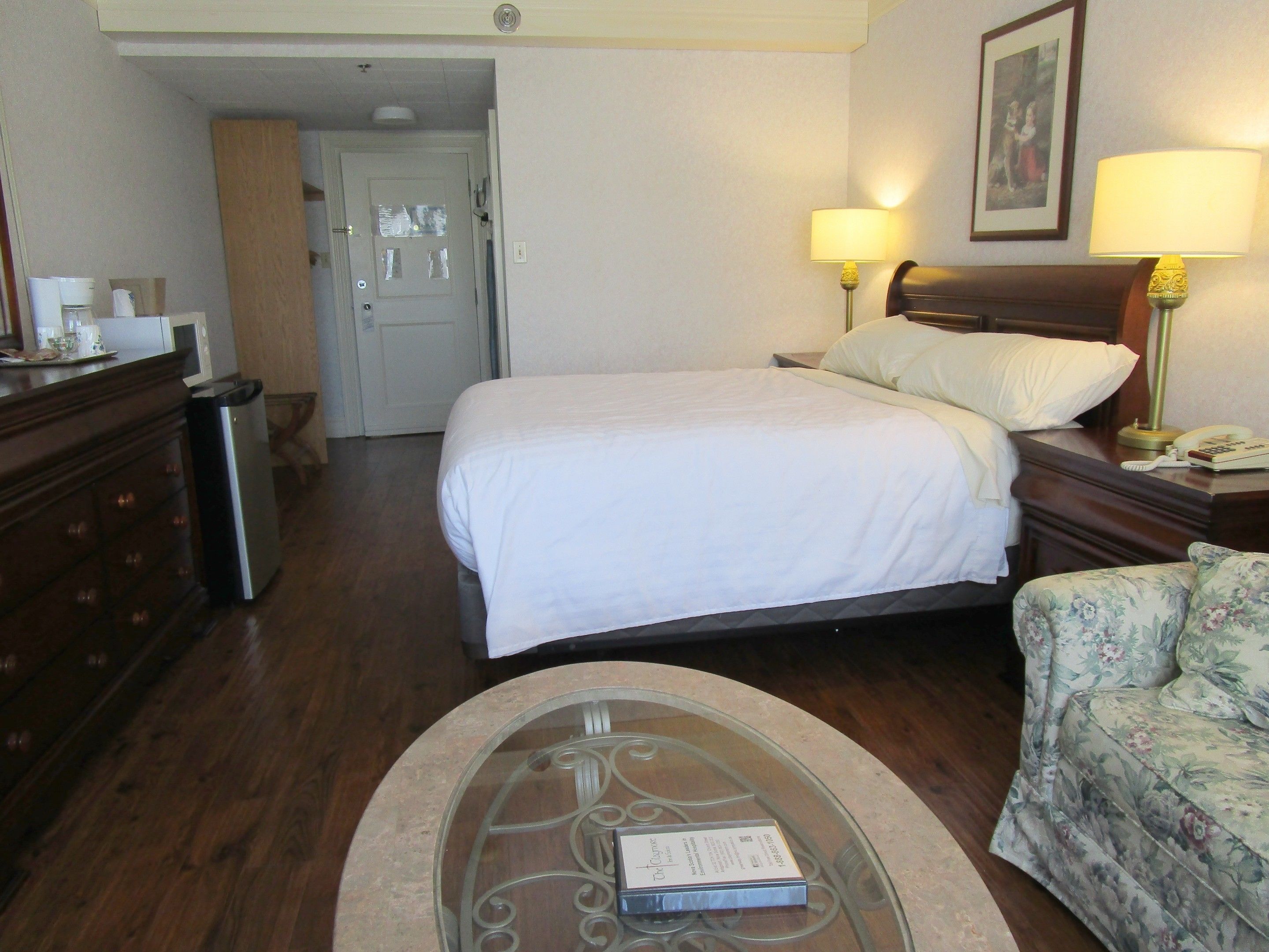 A hotel room with a bed, bedside lamps, a floral sofa, a glass coffee table with a book, a wooden dresser, and a door.