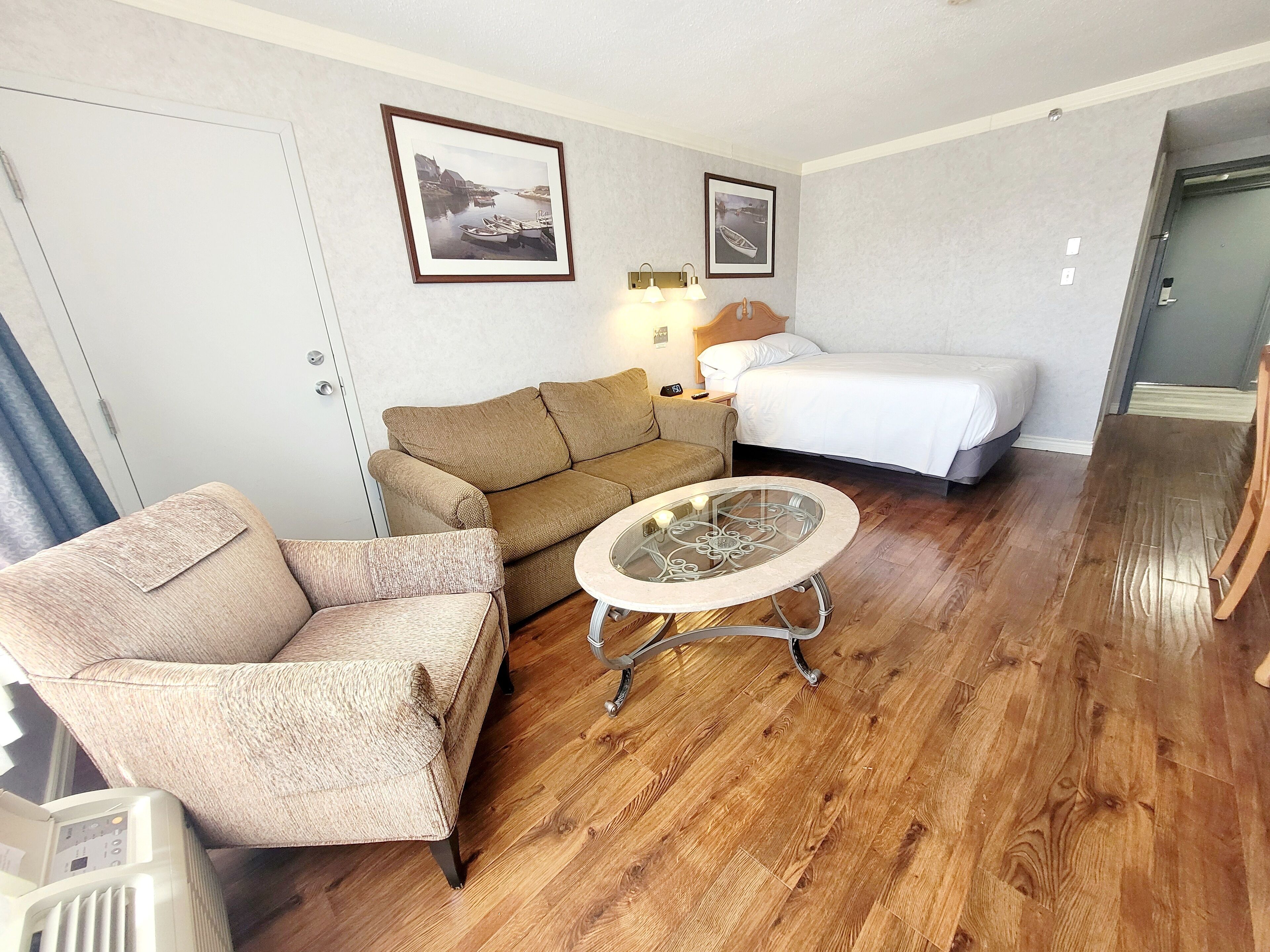 Une pièce avec un canapé marron, un fauteuil beige, une table basse en verre, un lit avec une literie blanche et deux tableaux encadrés accrochés au mur.