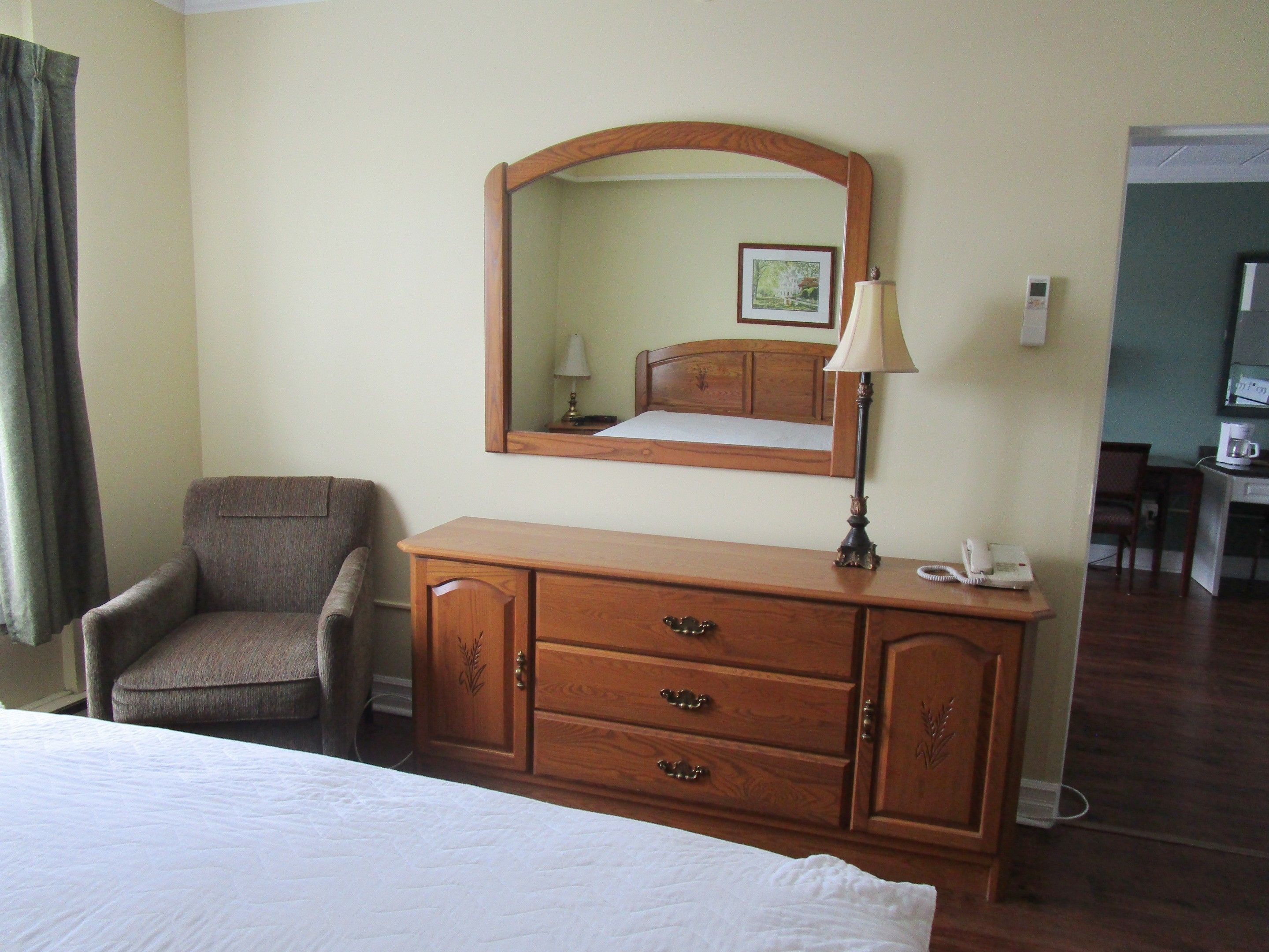 Habitación de hotel con cama, cómoda de madera, silla, escritorio con lámpara y espejo.
