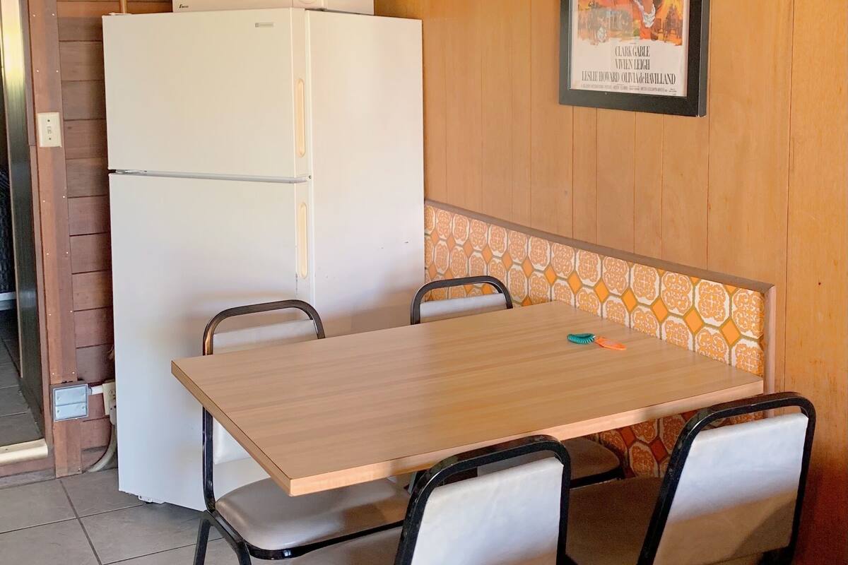 A small dining area with a wooden table and chairs, a white refrigerator, and a poster on the wall.
