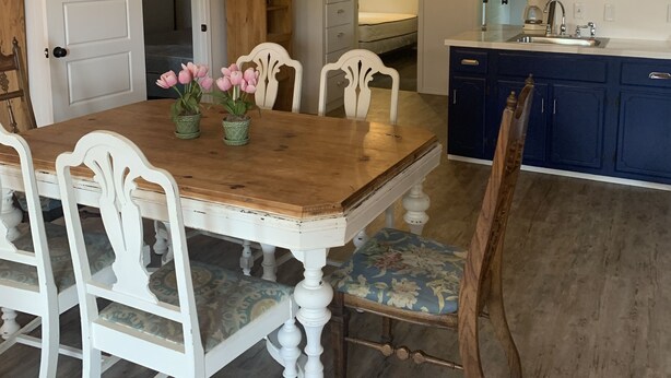 Un comedor con mesa de madera y sillas blancas, una cocina con armarios azules y un techo alto con vigas a la vista.
