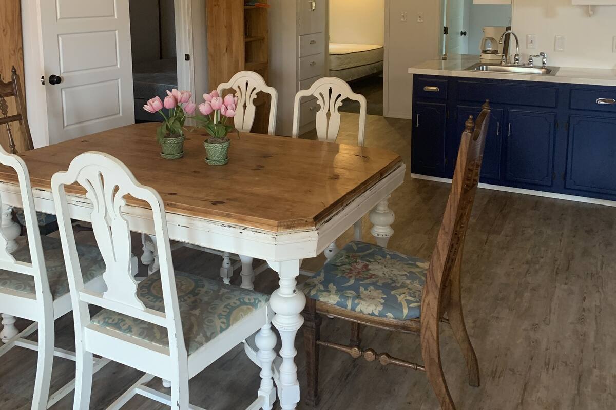 A dining area with a wooden table and white chairs, a kitchen with blue cabinets, and a high ceiling with exposed beams.