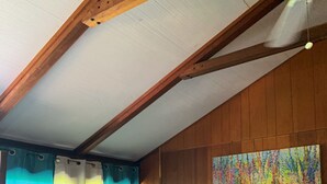Un dormitorio con cama, vigas de madera en el techo, ventilador de techo, un cuadro y una ventana con cortinas.