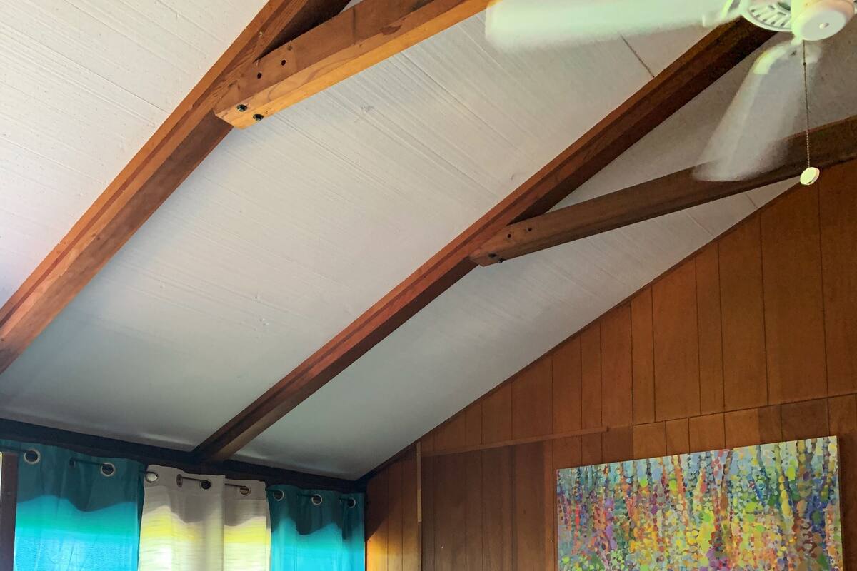 A bedroom with a bed, wooden ceiling beams, a ceiling fan, a painting, and a window with curtains.