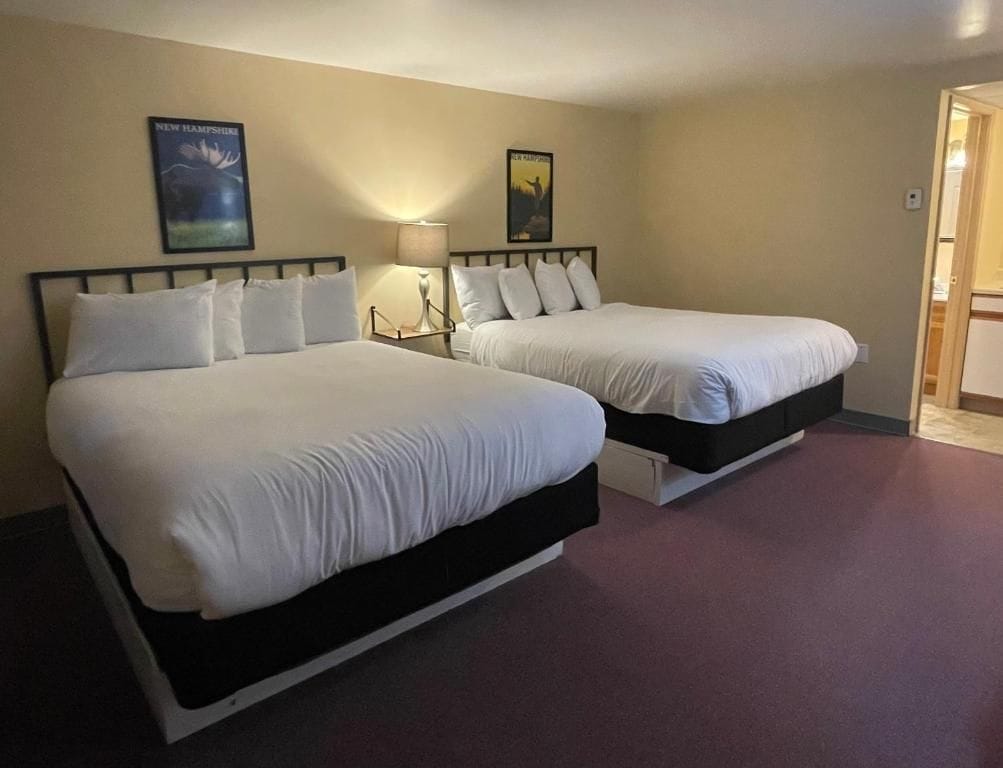 A hotel room with two beds, a nightstand with a lamp, and a door leading to a bathroom.