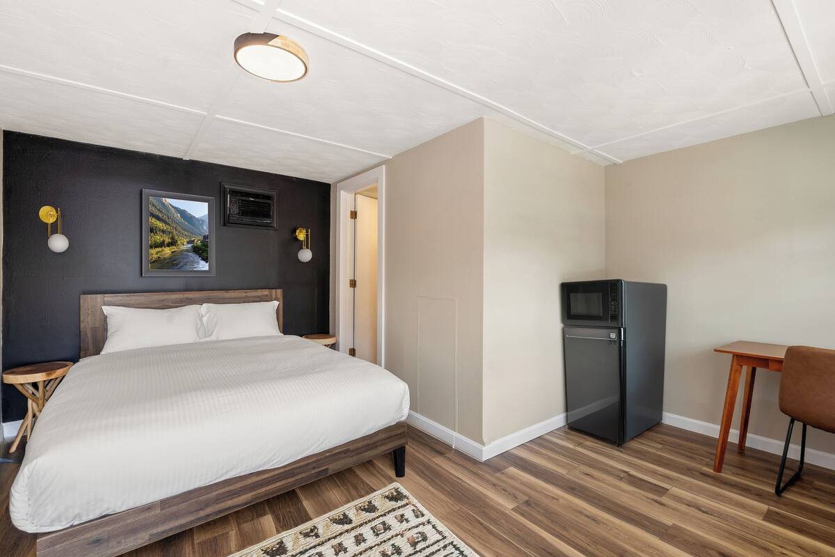 A bedroom with a bed, a small table, a chair, a refrigerator, and a picture on the wall.
