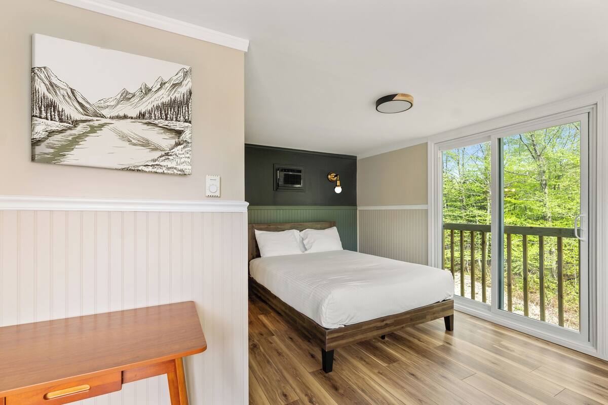 A bedroom with a wooden bed, a nightstand, a wall-mounted painting, a window with a view of greenery, and a ceiling light fixture.