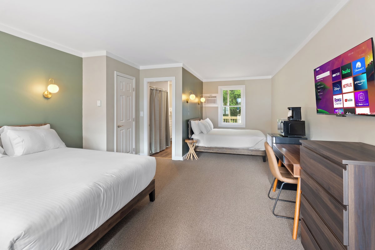 A hotel room with two beds, a desk, a TV displaying streaming services, and a view of the outdoors.