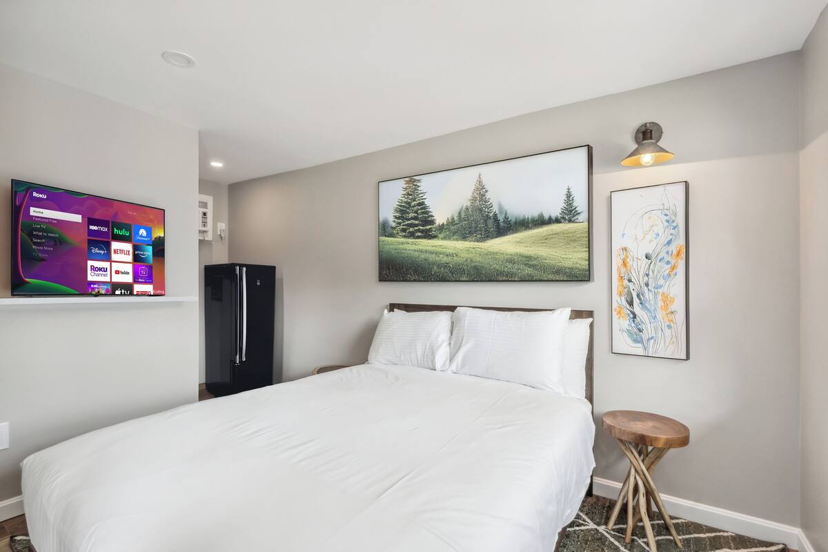 A hotel room with a bed, a flat-screen TV displaying streaming services, a refrigerator, and artwork on the walls.