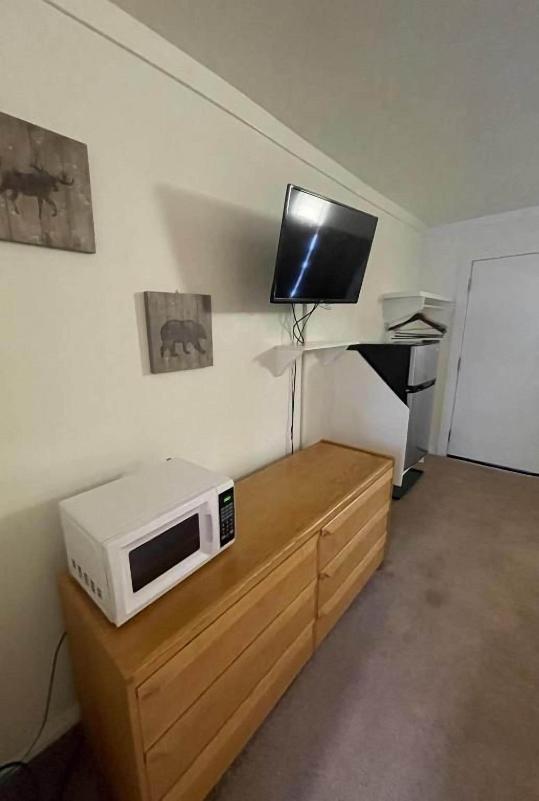 A room with a wooden dresser, a microwave, a flat-screen TV mounted on the wall, and a door in the background.