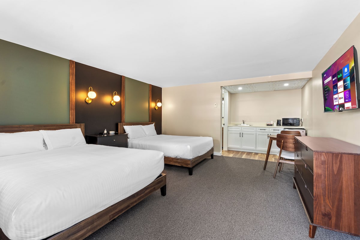 A hotel room with two beds, a desk, a chair, a TV, and a kitchenette.