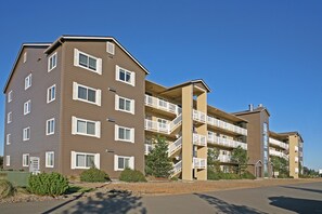 A multi-story apartment building with balconies, a parking lot, and a clear blue sky. - Outlook Condos at Oyhut Bay Resort (Ocean Shores)