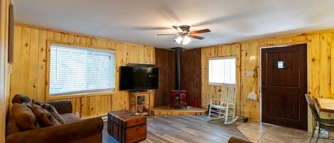 Una habitación acogedora de estilo cabaña con interior de madera, ventilador de techo, televisor, chimenea y una mecedora.