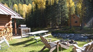 A rustic cabin with a picnic table and Adirondack chairs by a river. - Wolf Creek Ranch Ski Lodge (South Fork)