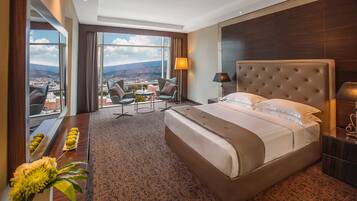 Una habitación de hotel con una cama grande, un escritorio, dos sillas, una mesita y vistas a la ciudad y montañas.