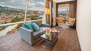 Hotelkamer met een groot raam en uitzicht over de stad, een bank, een stoel en een salontafel met een bloemenvaas.