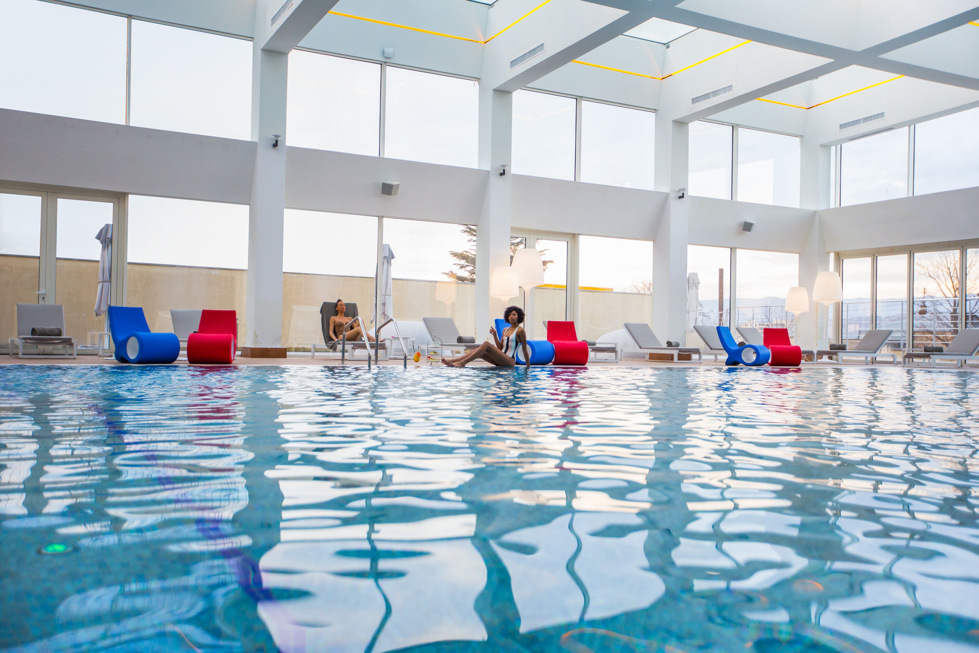 An indoor swimming pool with clear water, surrounded by lounge chairs and large windows.