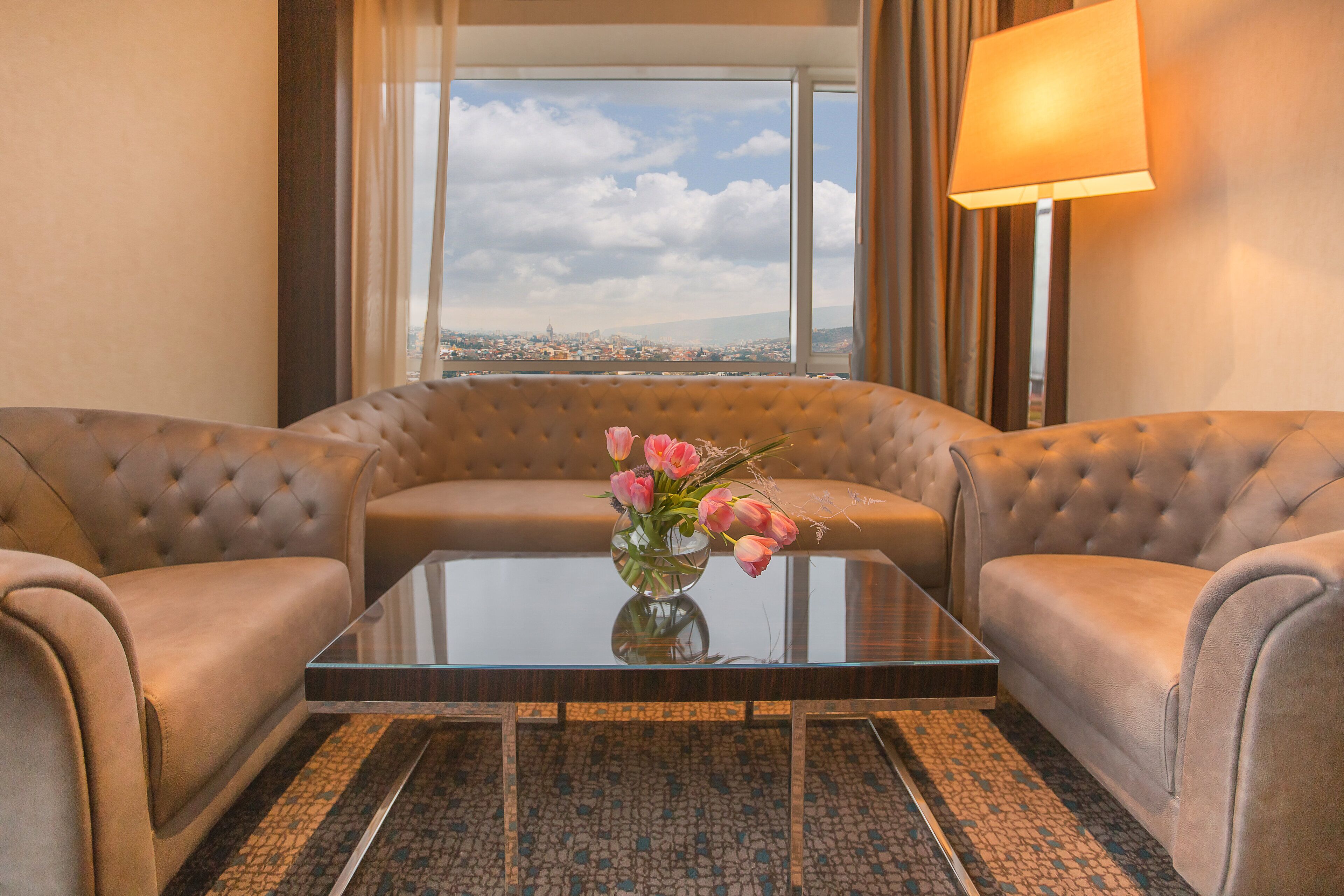A room with a sofa set, a glass coffee table with a vase of flowers, and a view of a cityscape through large windows.