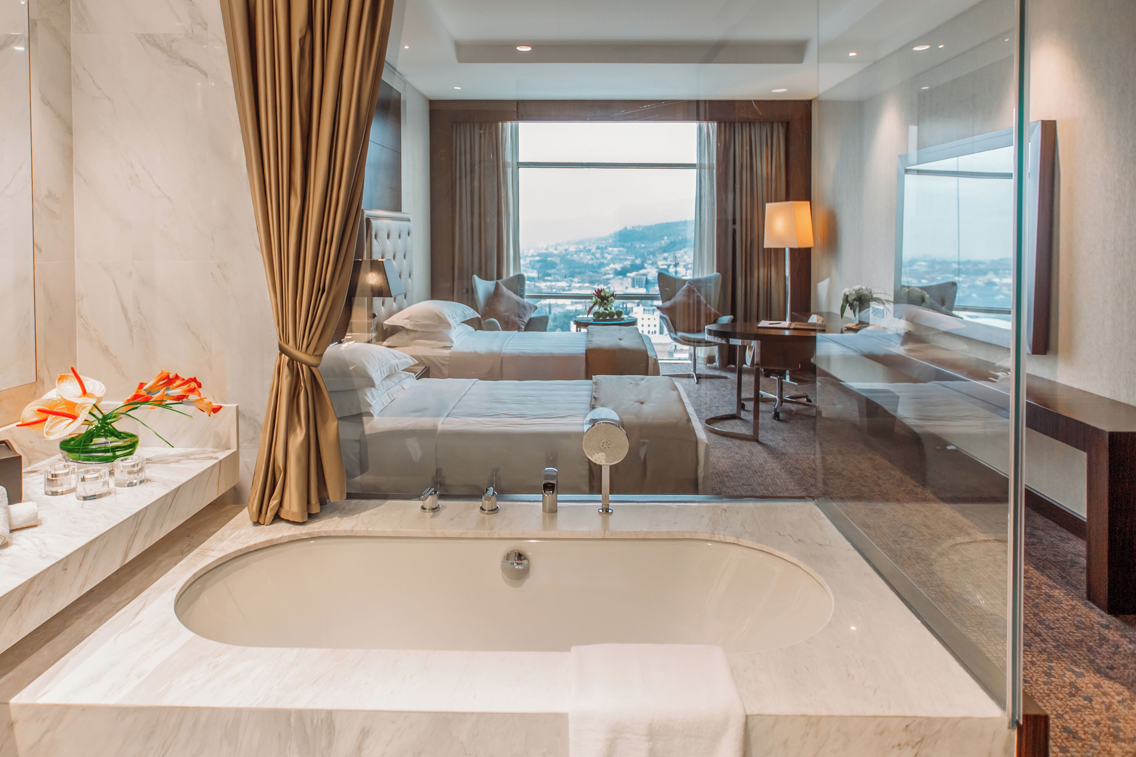 A modern hotel room with a large bathtub, a bed with a view, a desk, and a chair.