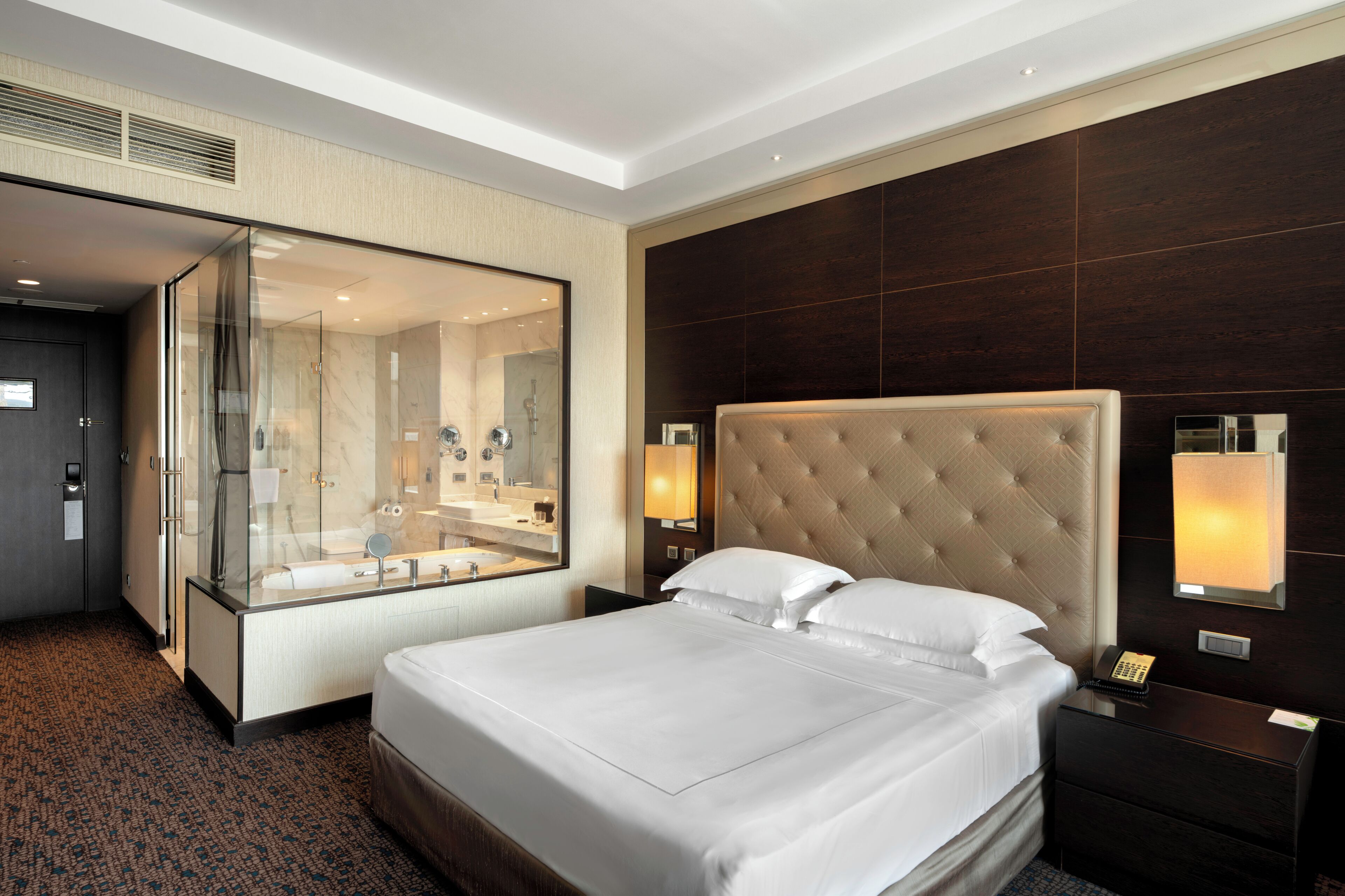 A hotel room with a large bed, a glass-enclosed bathroom, and a dark wood panel wall.