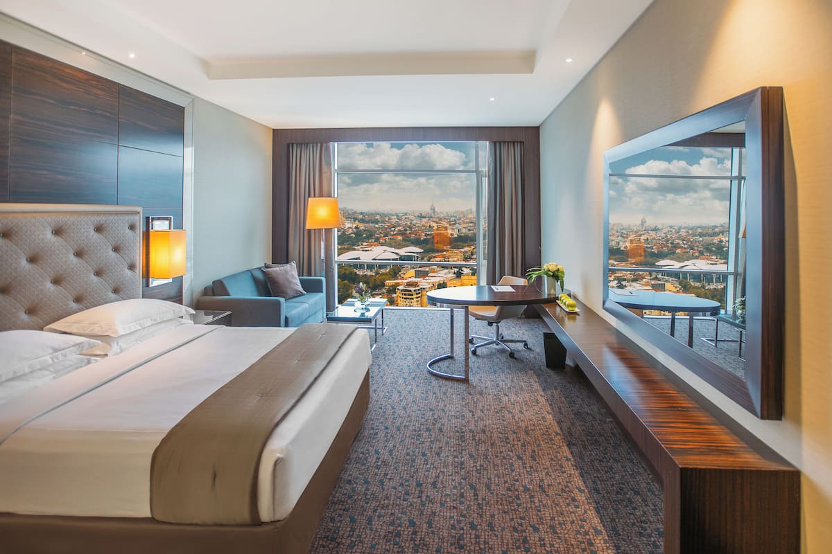 Ein Hotelzimmer mit einem großen Bett, einem Schreibtisch, einem Sessel und einer Aussicht auf die Stadt.