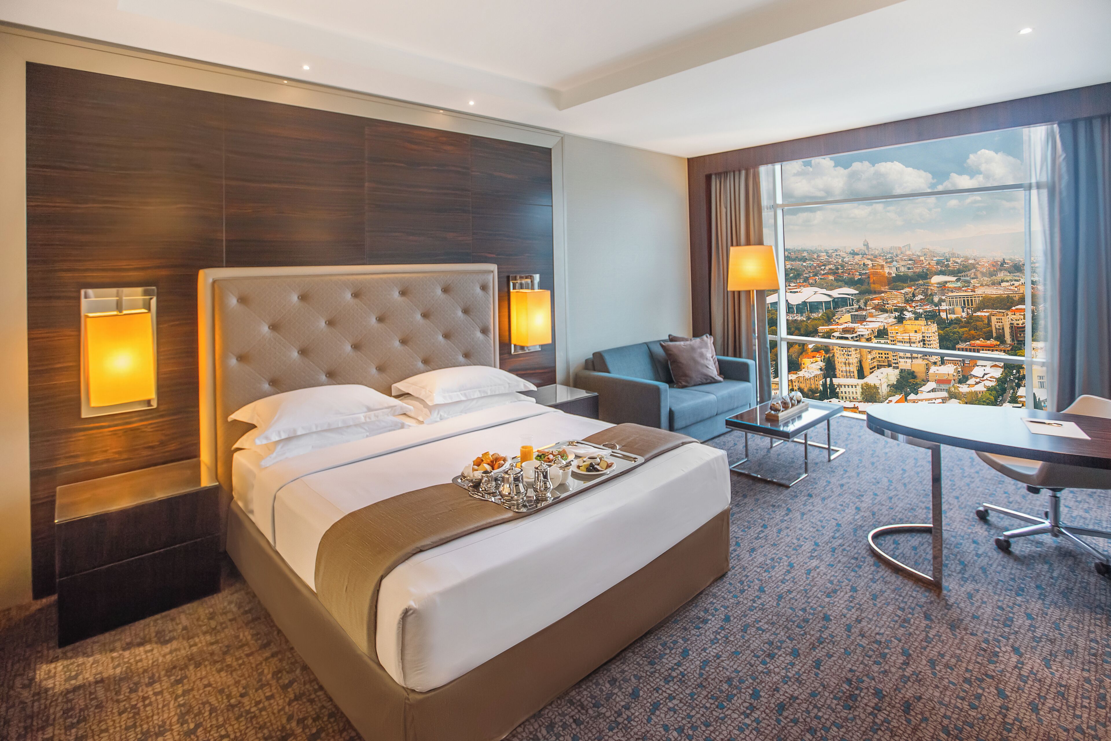 A hotel room with a large bed, a sofa, a desk, and a view of a cityscape.
