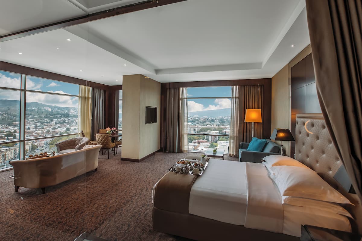 Ein Hotelzimmer mit einem großen Bett, einer Couch, einem kleinen Tisch und Blick auf eine Stadtlandschaft.