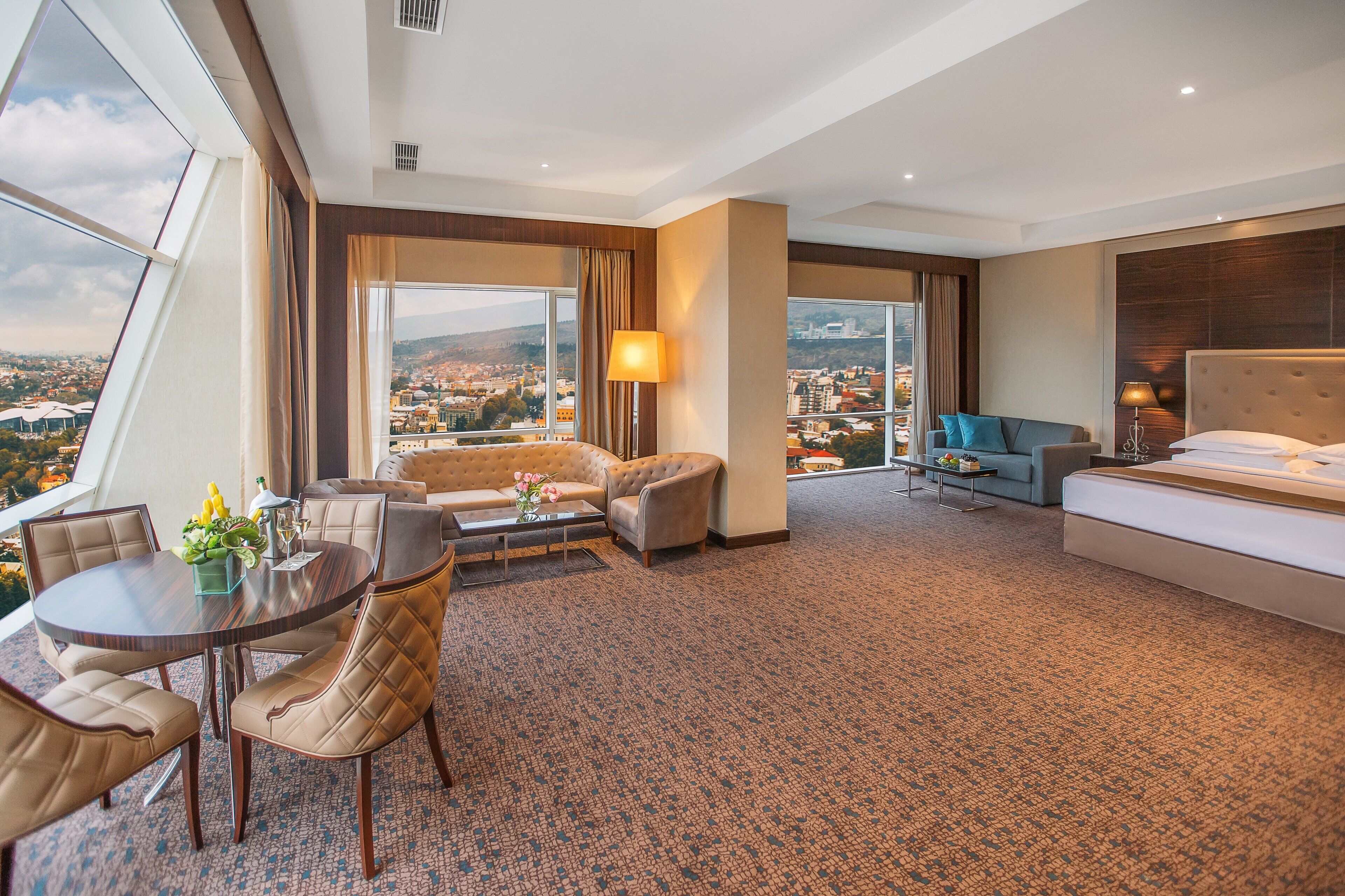A hotel room with a large bed, a sofa, a dining table with chairs, and a view of the city through large windows.