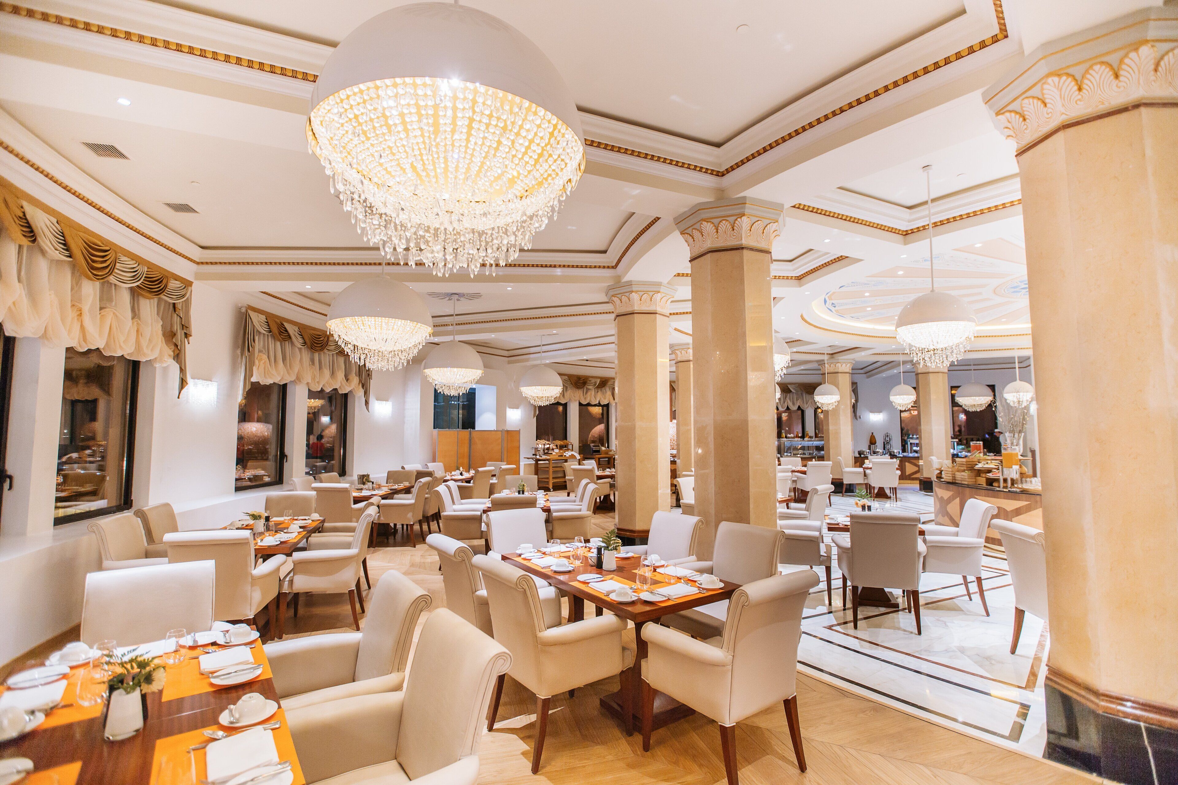 A spacious hotel dining area with multiple tables, elegant chandeliers, and classic decor.