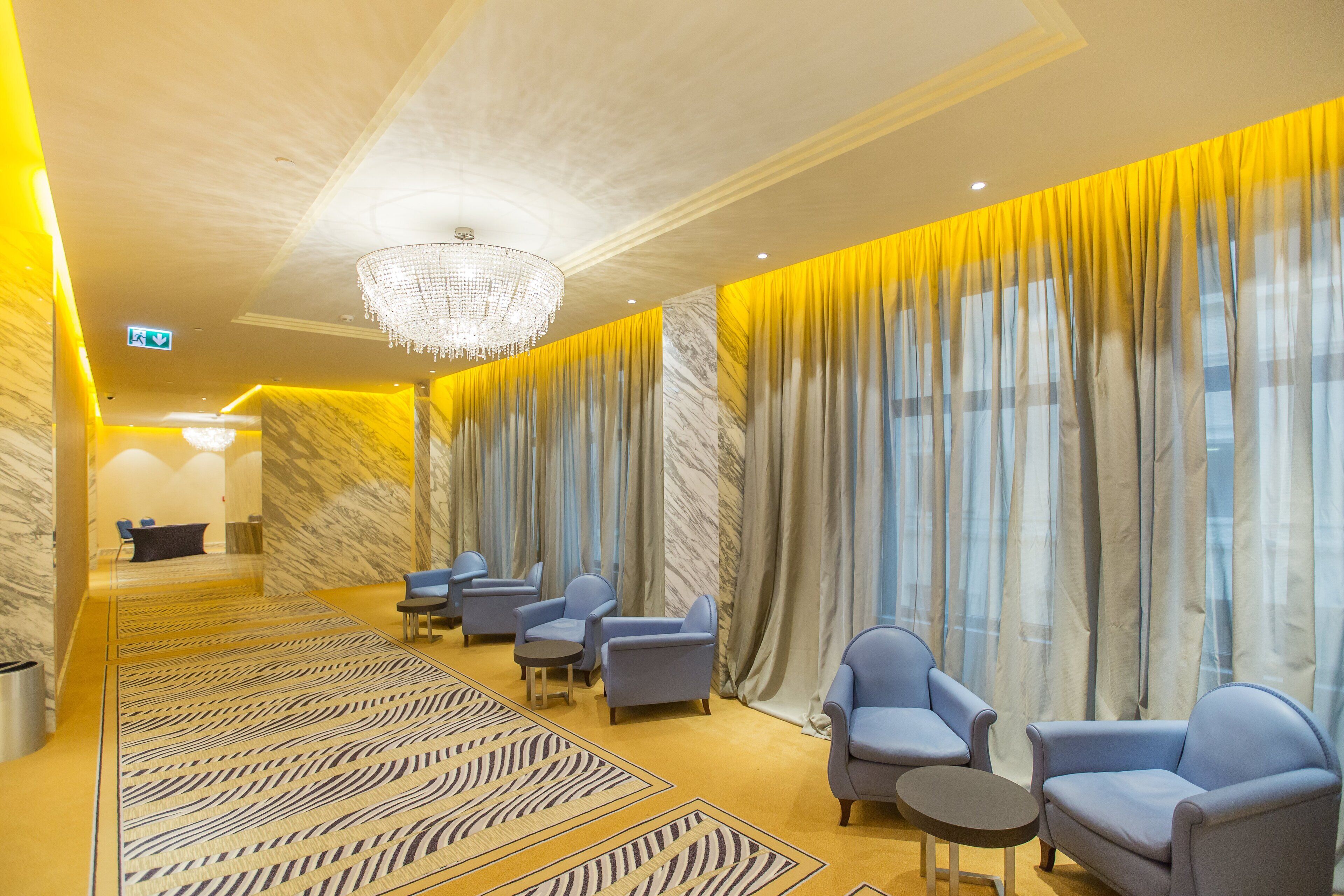 Un hall d’hôtel avec un comptoir de réception en marbre, des fauteuils bleus, une moquette à motifs et un lustre.